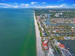 An aerial view of a coastal city with a long beachfront, dense residential area, and a marina.