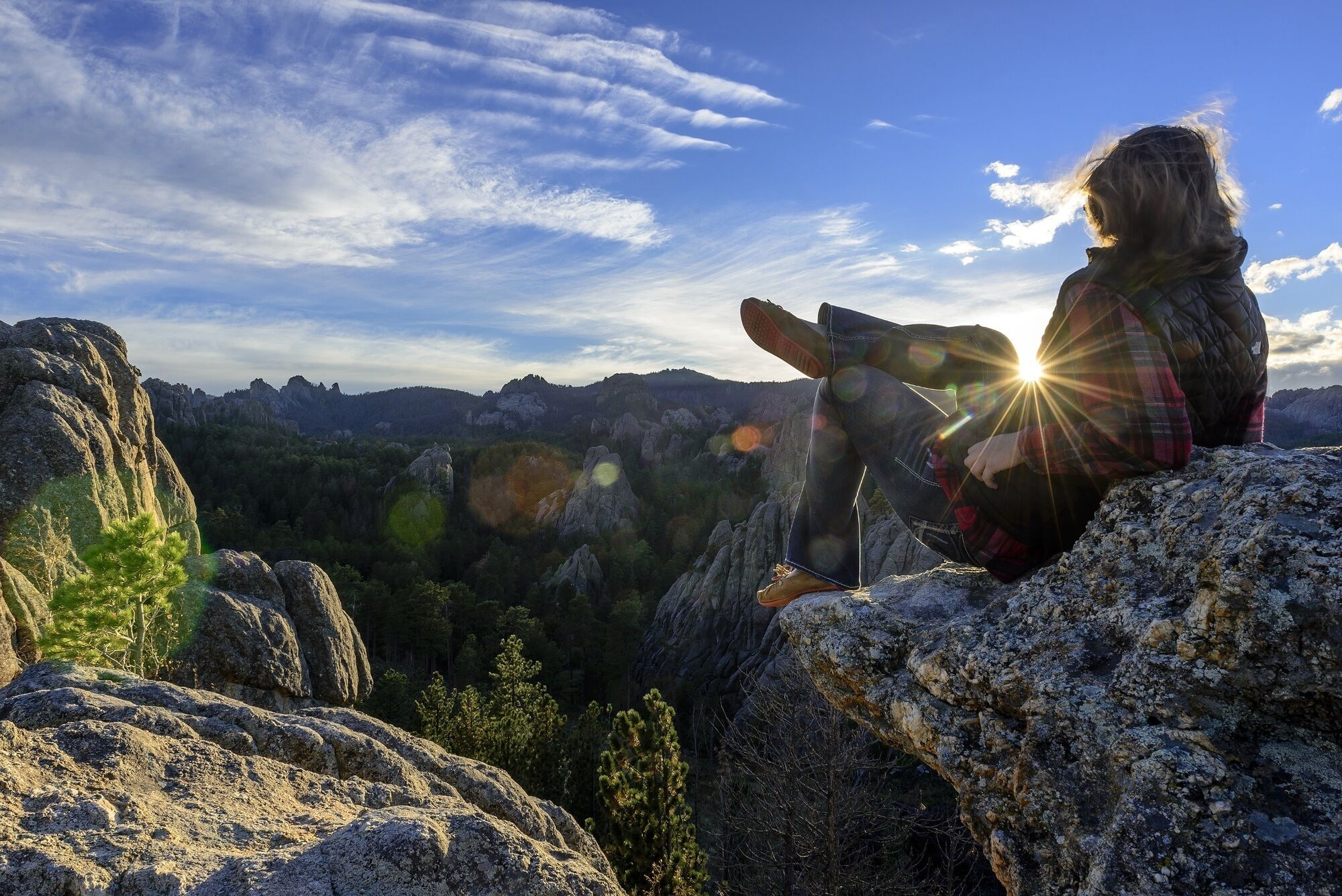 Une personne est assise sur un promontoire rocheux, d’où elle domine un paysage montagneux, tandis que le soleil brille à travers les nuages.