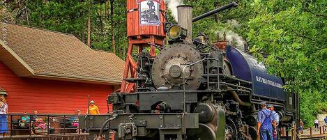 A steam locomotive with a red and blue color scheme, a chimney emitting steam, and a small building with a sign reading "1880 TRAIN."