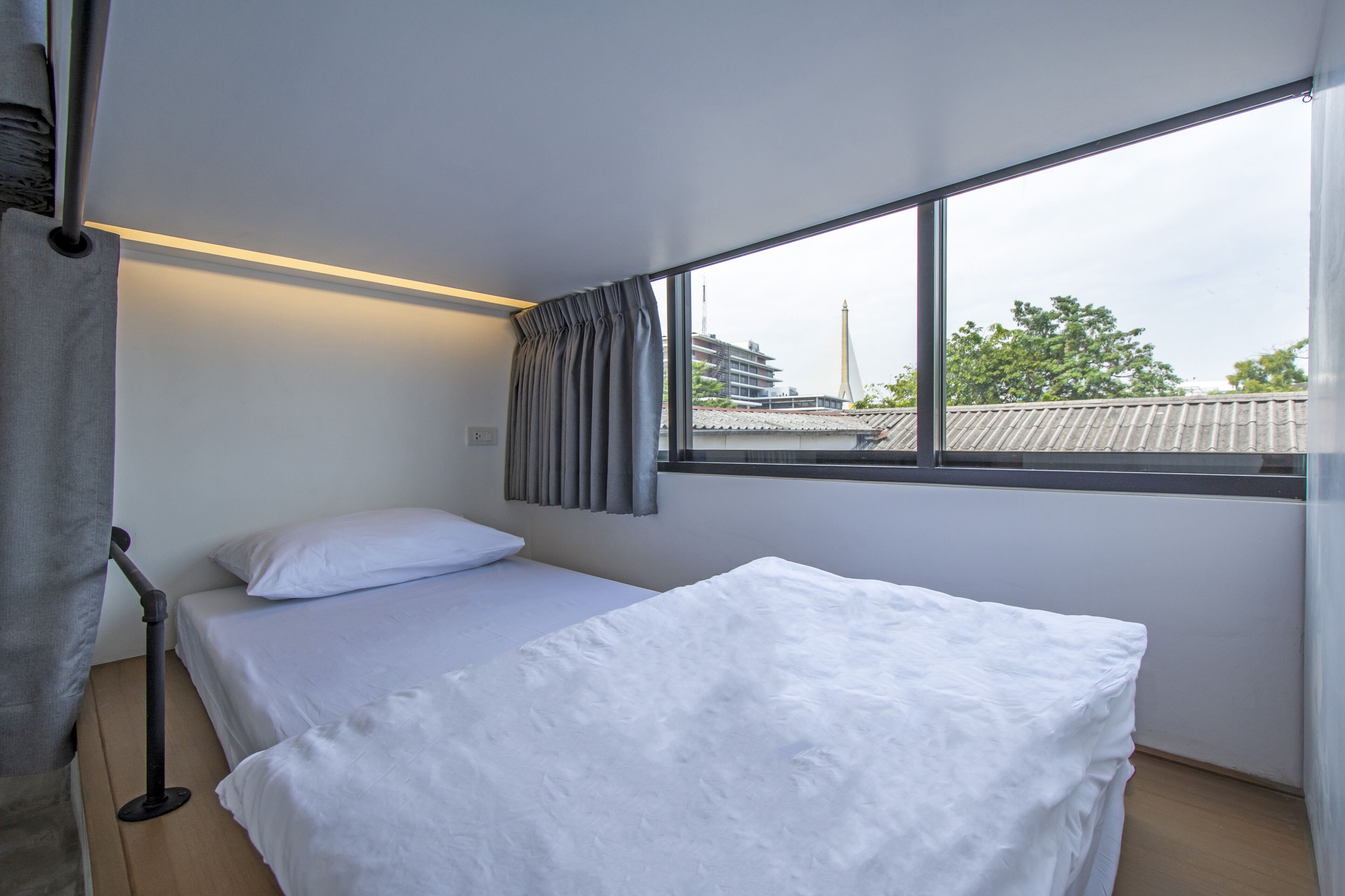 A single bed with white bedding in a room with a large window, curtains, and a visible cityscape outside.