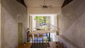 Un pasillo estrecho con ventilador de techo, una mesa de madera con sillas, y vista a una escalera y follaje exterior.