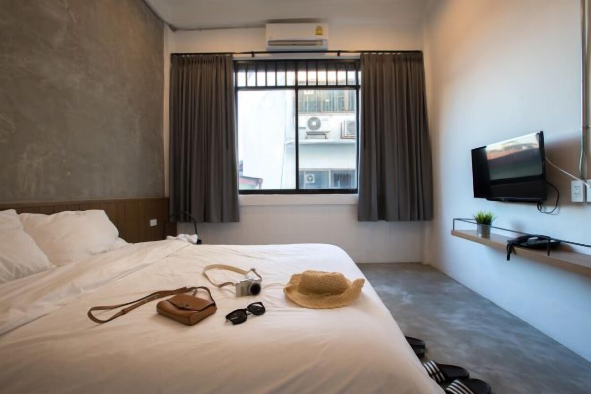 A hotel room with a bed, a TV mounted on the wall, a window with curtains, and a small shelf with a plant and a phone.
