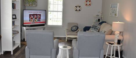 Een woonkamer met een televisie, twee fauteuils, een salontafel, een boekenkast en een raam met jaloezieën.