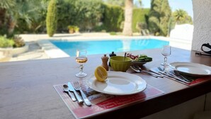 A dining table with a view of a swimming pool, a glass of white wine, a bowl of salad, and a lemon on a red placemat.