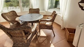 A sunlit patio with wicker furniture, a round table, and large windows with white curtains.