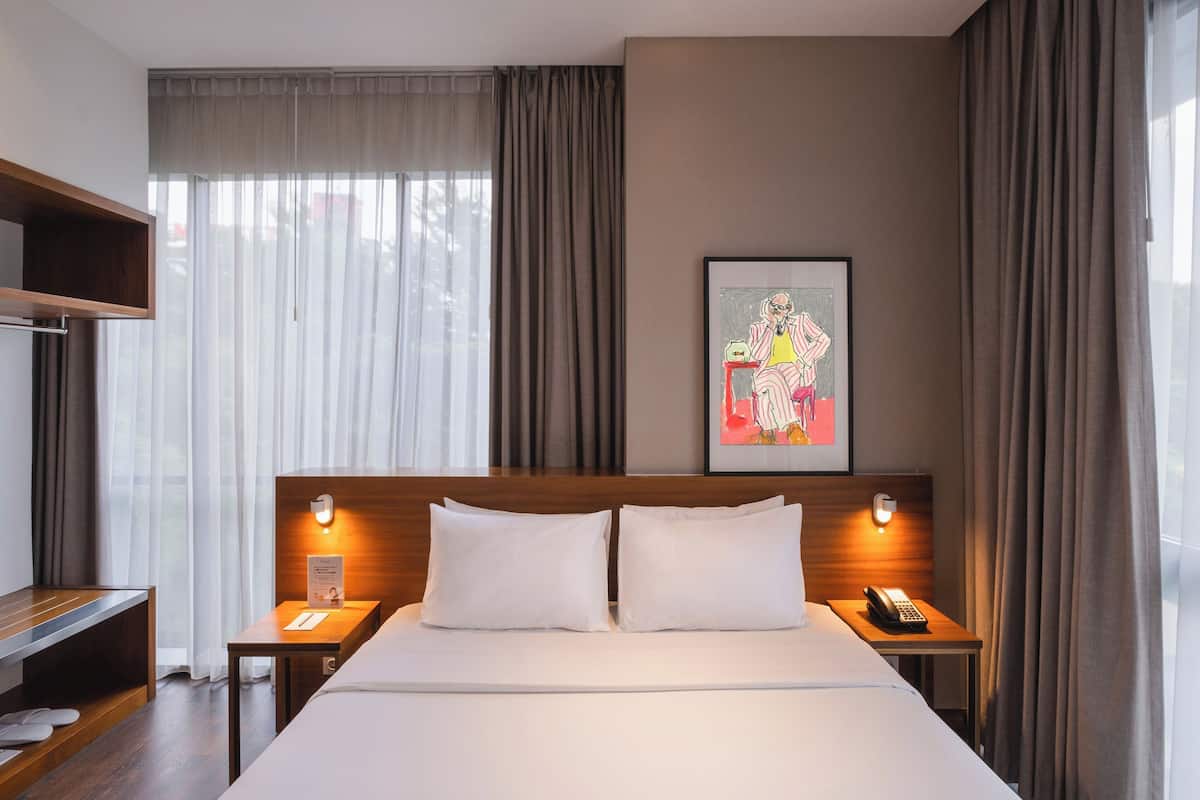 Ein Hotelzimmer mit einem großen Bett, Nachttischen, einer hölzernen Bank, einem gerahmten Bild an der Wand und einem Fenster mit Vorhängen.