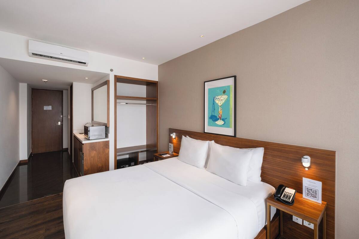 Ein Hotelzimmer mit einem großen Bett, einem Nachttisch mit Telefon und einem QR-Code, einem weiteren Nachttisch mit Lampe, einem Kleiderschrank und einem Bild an der Wand.