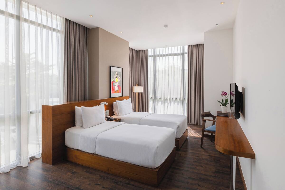 Ein Hotelzimmer mit zwei Betten, einem Schreibtisch, einem Stuhl, einem Fernseher und großen Fenstern mit Vorhängen.