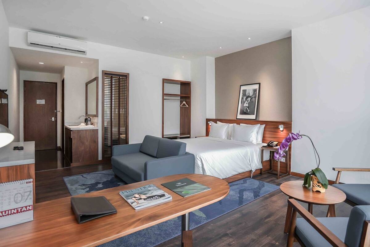 Ein Hotelzimmer mit einem Bett, zwei Sofas, einem Schreibtisch mit Lampe, einem kleinen Tisch und einem durch eine offene Tür einsehbaren Badezimmer.