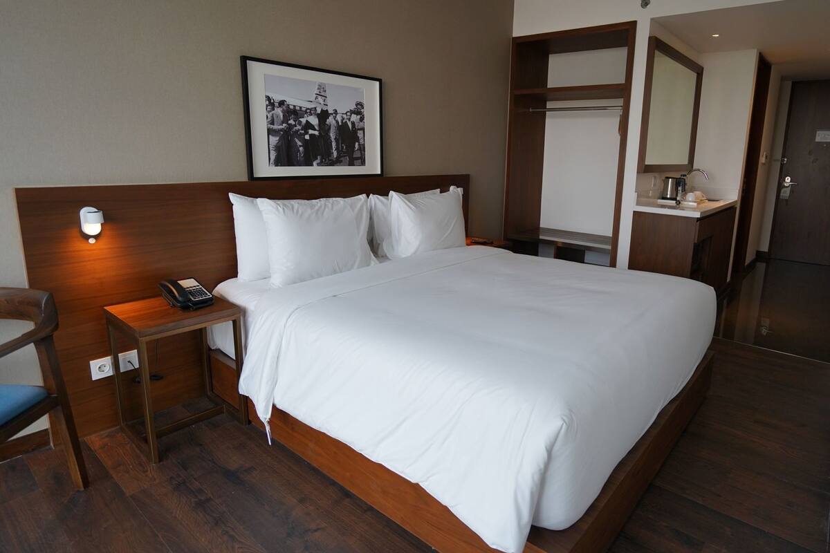 Ein Hotelzimmer mit einem großen Bett, einem Nachttisch mit Telefon, einem Spiegel und einem gerahmten Foto an der Wand.