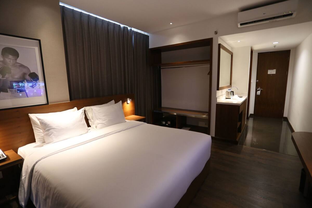 Ein Hotelzimmer mit einem großen Bett, einem Nachttisch, einem Kleiderschrank, einem Schreibtisch, einem Spiegel und einer Klimaanlage.
