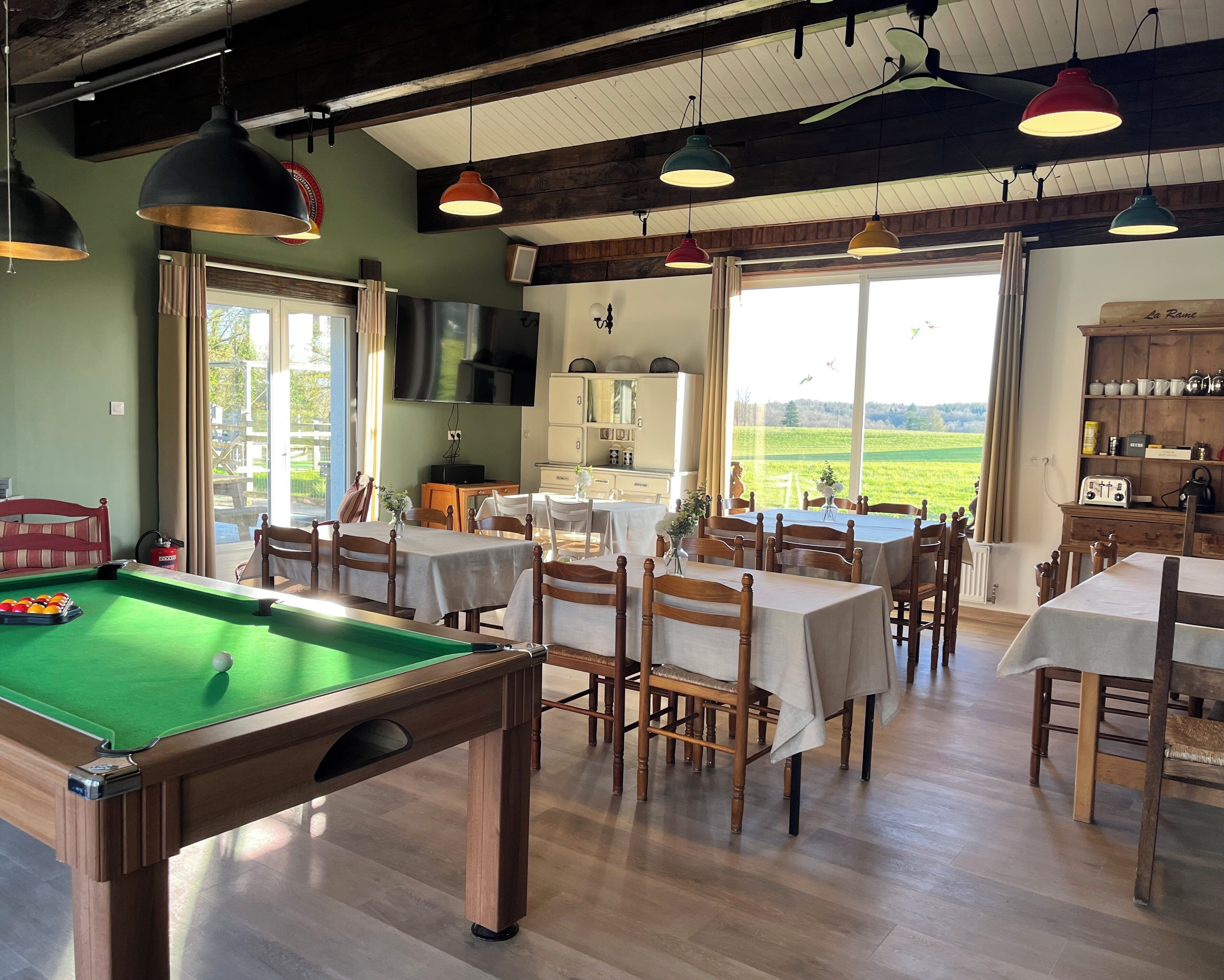 Une pièce dotée d’une table de billard, de tables et de chaises, et offrant une vue sur un champ verdoyant grâce à de grandes fenêtres.