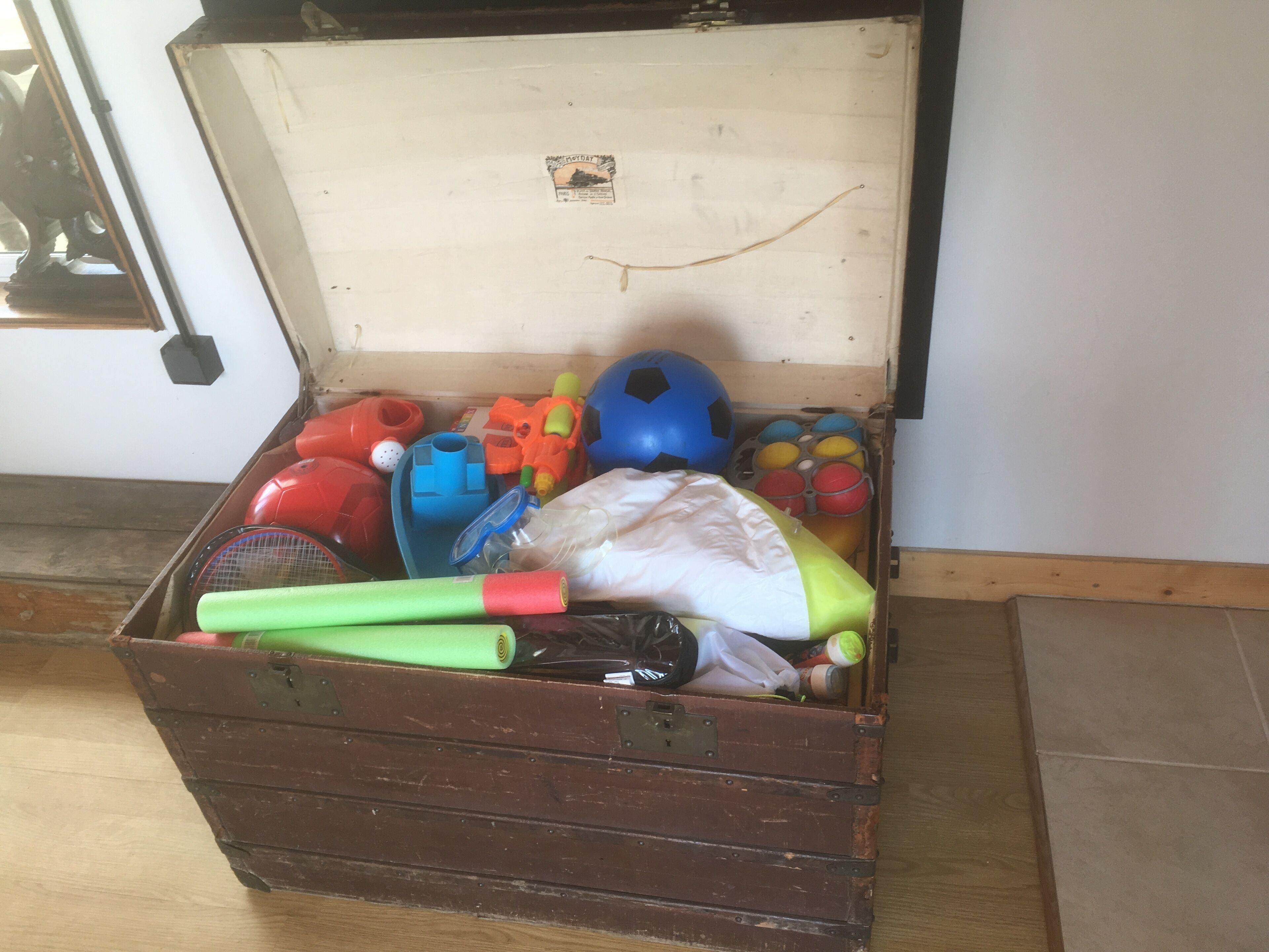 A wooden chest filled with various sports equipment including a soccer ball, badminton rackets, and a tennis racket.