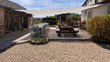 A rustic outdoor dining area with a stone table and benches, surrounded by stone walls and a gravel ground.