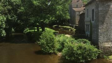 Edifici històric amb parets de pedra i un teulat tradicional, situat al costat d'un riu tranquil i vegetació abundant.