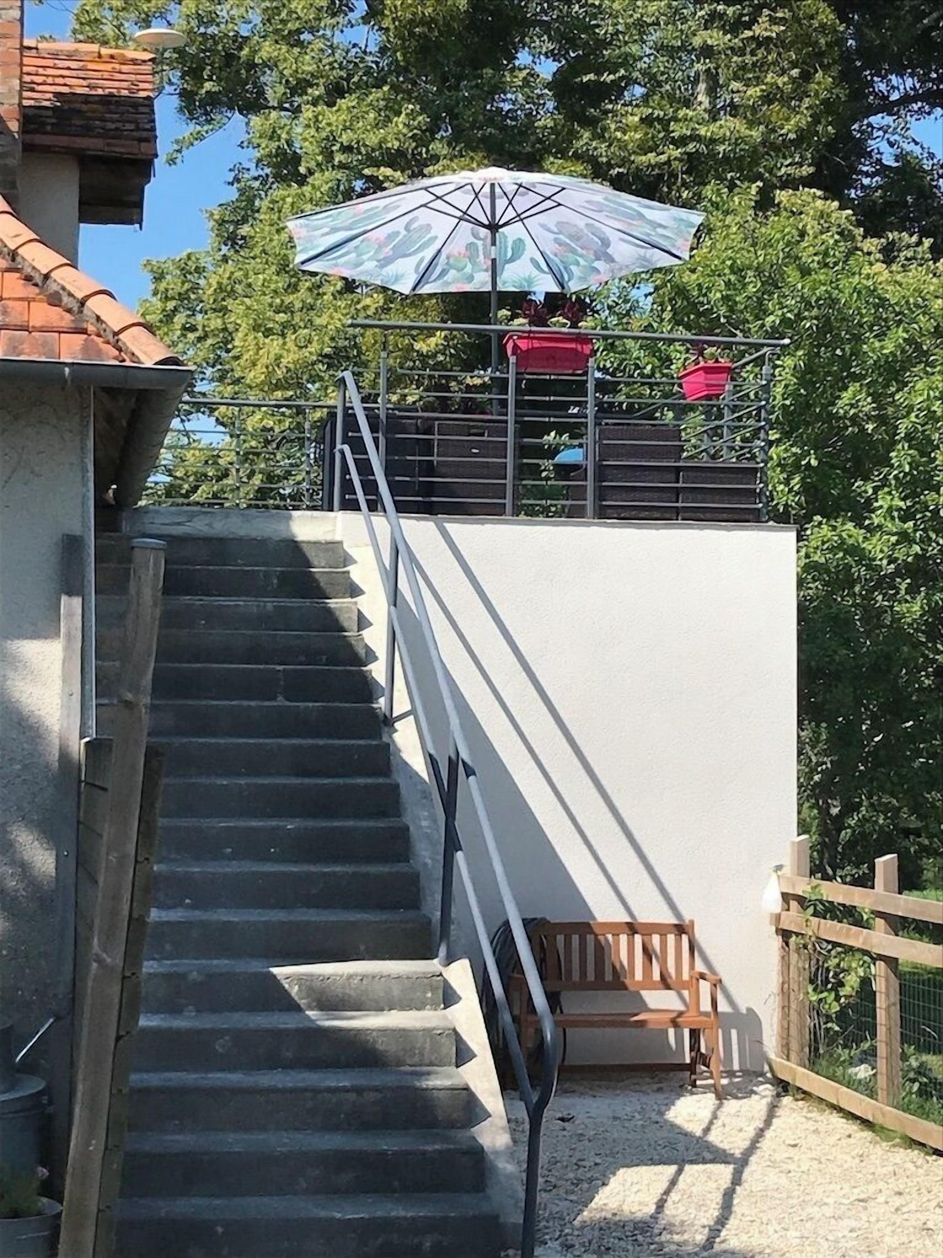 Un escalier menant à un balcon agrémenté d’un parasol coloré, d’un banc en bois et d’une clôture.