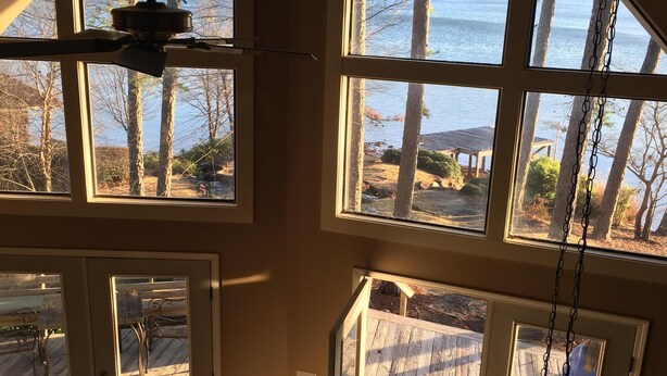 Una habitación con amplios ventanales que dan a un lago, un ventilador de techo y vistas a una terraza de madera.