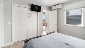 Ein Schlafzimmer mit Bett, einem an der Wand montierten Fernseher, einem Fenster mit Jalousien und einer Tür zu einem anderen Raum.
