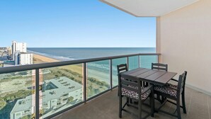 En balkon med bord og stole, der vender ud mod en strand og et hav.