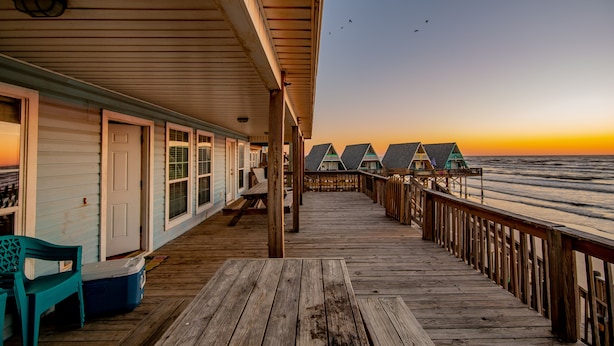 Un muelle de madera que lleva a cabañas frente a la playa, con vista al atardecer.