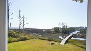 Blick von einer Veranda auf eine Wiese mit einer Holzbrücke über dem Wasser, Bäume und ein entferntes Gebäude.