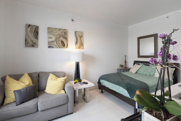 Un dormitorio moderno con un sofá gris, una mesita de noche con lámpara, una cama con ropa de cama verde, un espejo y una orquídea en maceta.