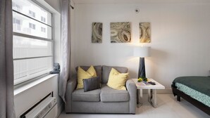 Een kamer met een grijze bank, een klein tafeltje met een lamp, een bed met een groene spreideken, en drie abstracte schilderijen aan de muur.