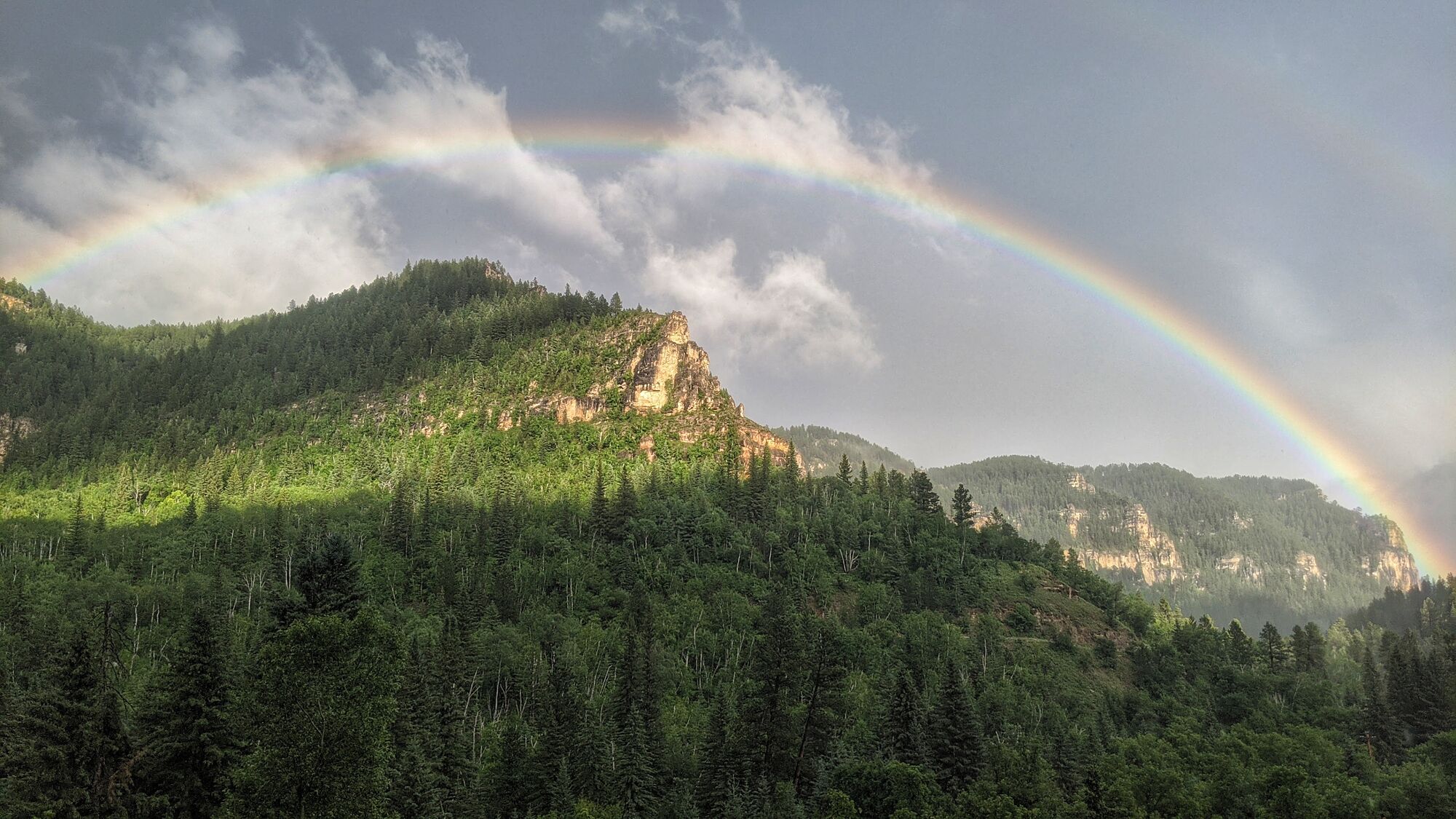 A forested mountain landscape with a rainbow arching over a rocky cliff.