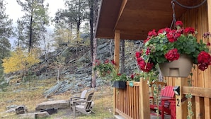 A wooden deck with a hanging planter of red flowers, a red Adirondack chair, and a stone fireplace. - Spectacular setting, amenities, and centrally located to Mt. Rushmore and Custer State Park, and hiking. Wi-Fi. Excellent cell service (Verizon & ATT).A favorite cabin for honeymoons and anniversaries Private campfire. Off-season discounts. No extra (Rapid City)