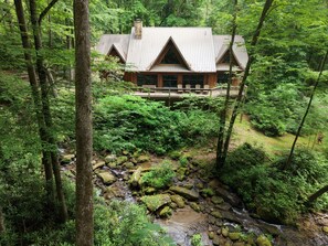 Une cabane dotée d’une terrasse, entourée d’une forêt dense et bordée d’un ruisseau.