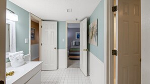 Um corredor com uma porta branca, um banheiro com pia branca e uma toalha, e um quarto com cama e um quadro na parede.