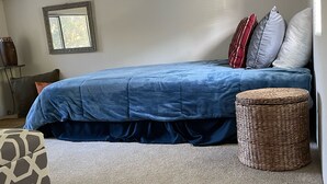 A bedroom with a bed, a small table, a chair, a window, and a ceiling vent.