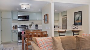 A modern kitchen with stainless steel appliances, a dining area with wooden chairs, and a living room with a beige sofa.