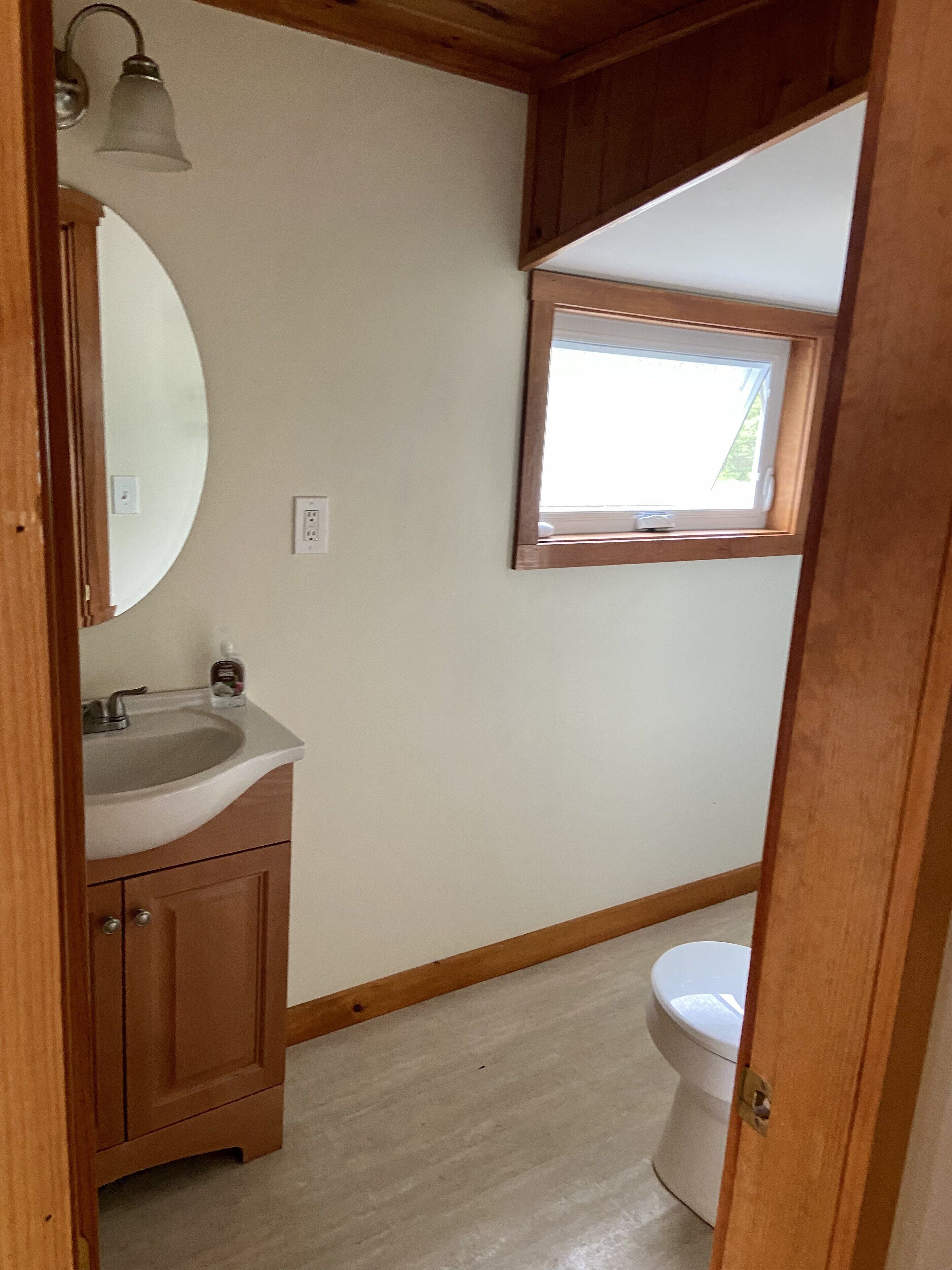 Ein kleines Badezimmer mit einem Holzbadschränken, einem runden Spiegel, einem Waschbecken und einer Toilette. Über der Toilette befindet sich ein Fenster.