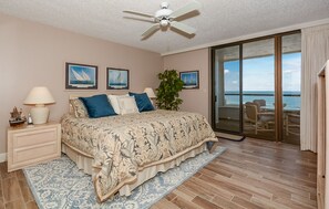 A bedroom with a bed, a nightstand, a lamp, a plant, and a view of the ocean through a sliding door.