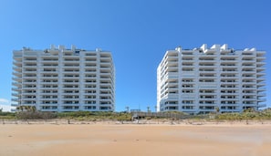Two high-rise buildings with multiple balconies, situated on a sandy beach with sparse vegetation.