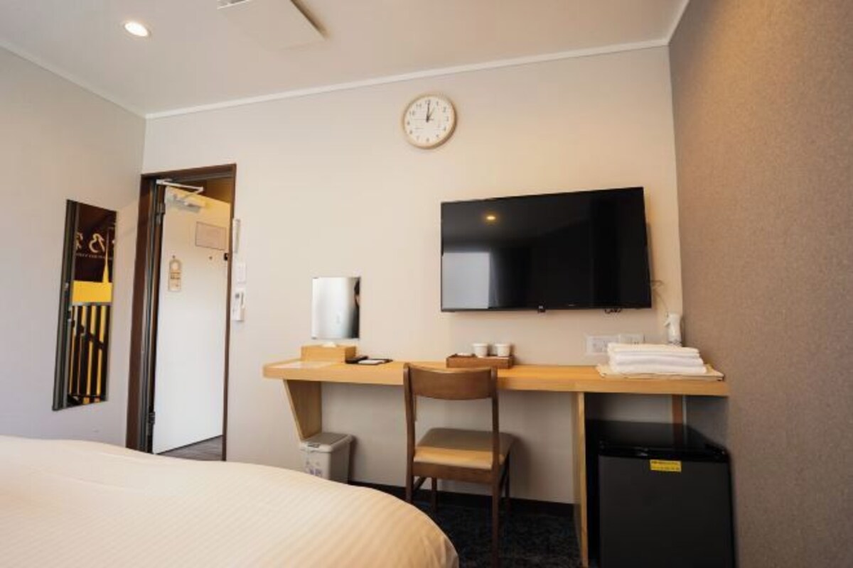 Une chambre d’hôtel comprenant un lit, un bureau avec une chaise, une télévision, une horloge et un petit réfrigérateur.