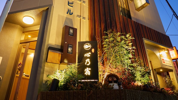 A building with signage indicating it is a hotel. There is a large illuminated sign with Japanese characters, a smaller sign with English text, and a decorative plant in front. - 心乃宿 Kokoro No Yado (Nagoya)
