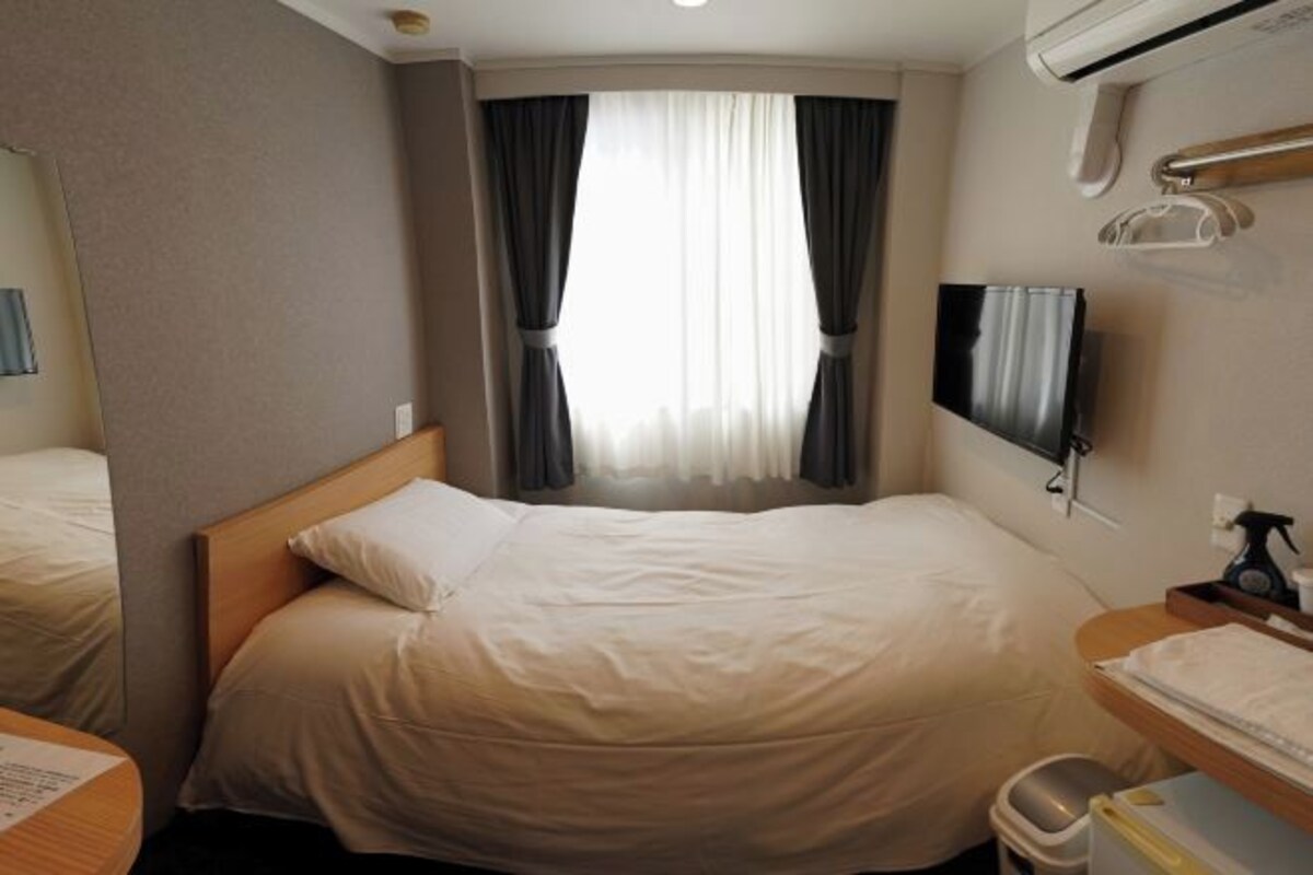 Une chambre d’hôtel comprenant un lit, une télévision, un miroir, une fenêtre avec des rideaux et une petite table avec une poubelle.