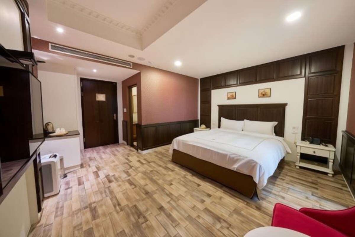 A bedroom with a large bed, a bedside table, a red chair, and a wooden floor.