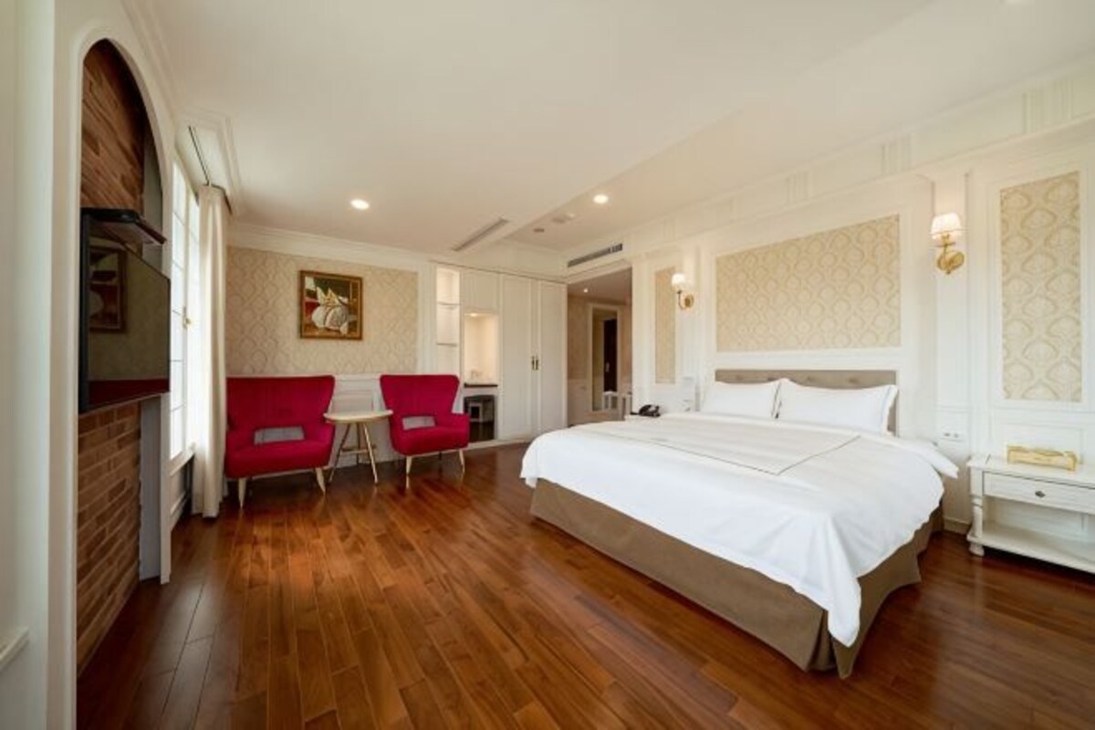A hotel room with a large bed, two red chairs, a small table, and a white nightstand.