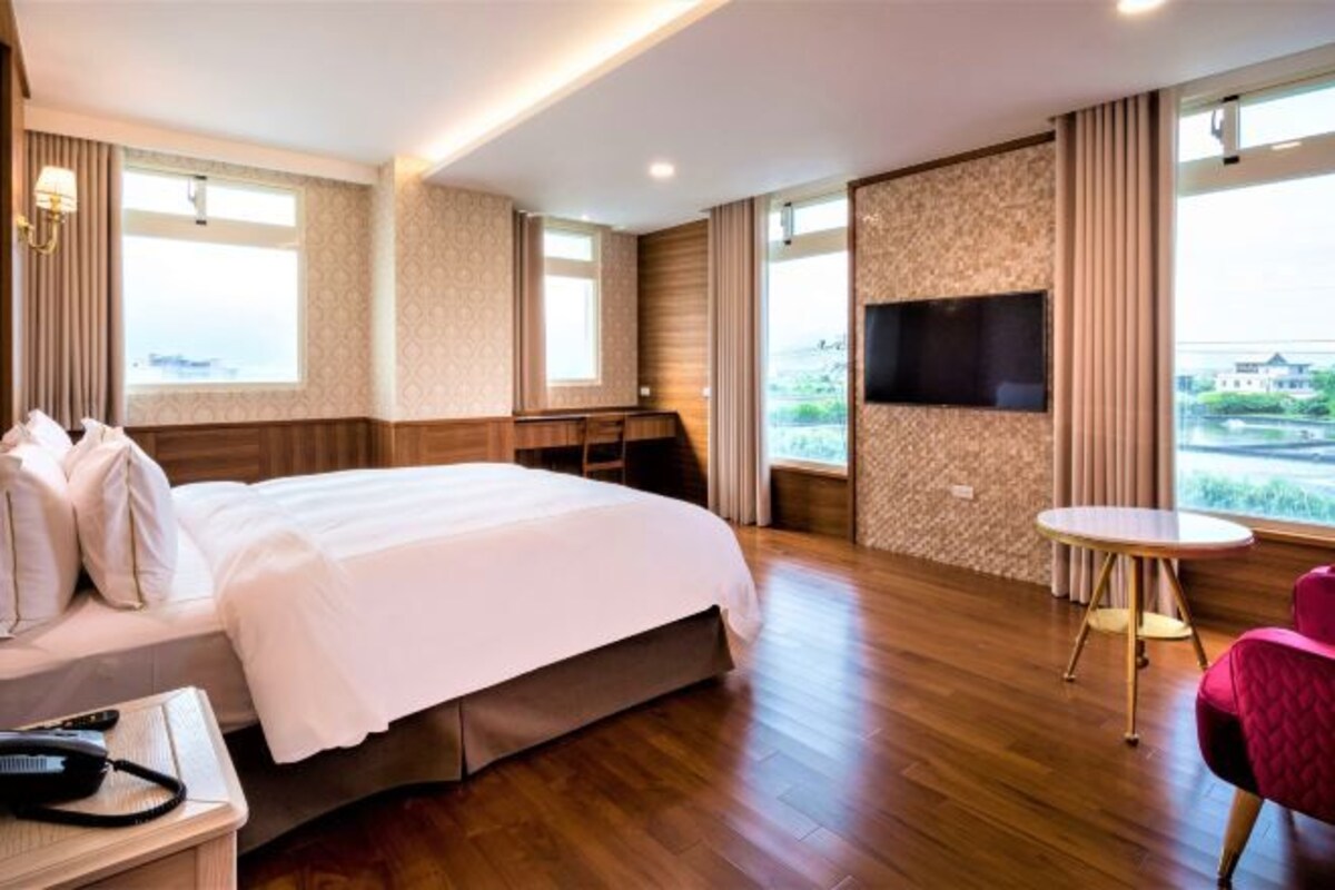Quarto de hotel com uma cama grande, uma escrivaninha com cadeira, uma mesinha redonda e uma televisão fixada na parede.