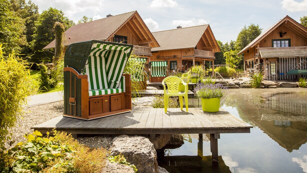 A wooden cabin with a deck overlooking a pond, surrounded by greenery. - Landgasthof Hessenmühle (Grossenlueder)