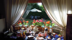 A table set with food and drinks, a window with curtains, and a view of a garden with flowers and a chair outside. - Podere La Piana (Fivizzano)