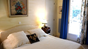 Una cama bien tendida con una colcha blanca acolchada y almohadones decorativos, una lámpara de noche y un cuadro con flores en la pared.