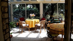 Un patio soleado con techo de vidrio, mobiliario de mimbre y una mesa con mantel amarillo.