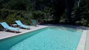 Una piscina rectangular con agua azul cristalina, rodeada por un solárium con baldosas. Hay tres sillas de descanso con cojines azules y una pequeña mesa. El área está a la sombra de árboles y tiene una planta en maceta.