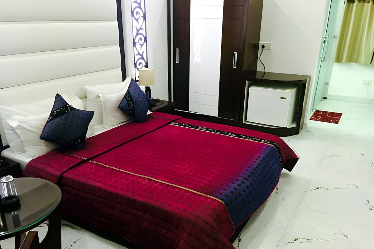 A hotel room with a bed, pillows, a small table with a lamp, a nightstand, and a mini-fridge.