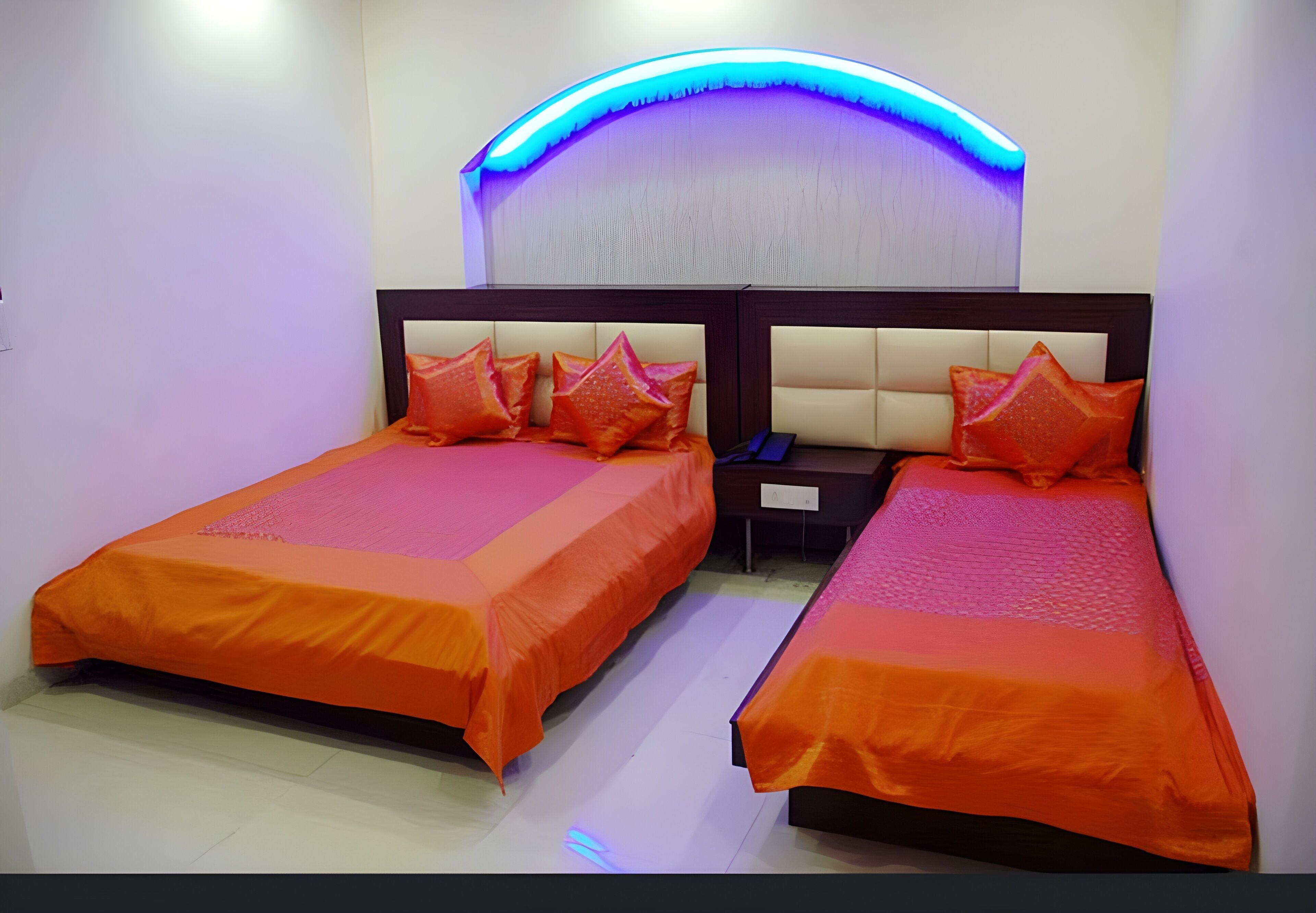 Een hotelkamer met twee bedden, elk met een oranje sprei en bijpassende kussens. Daartussen bevindt zich een nachtkastje, en boven is het plafond blauw verlicht.