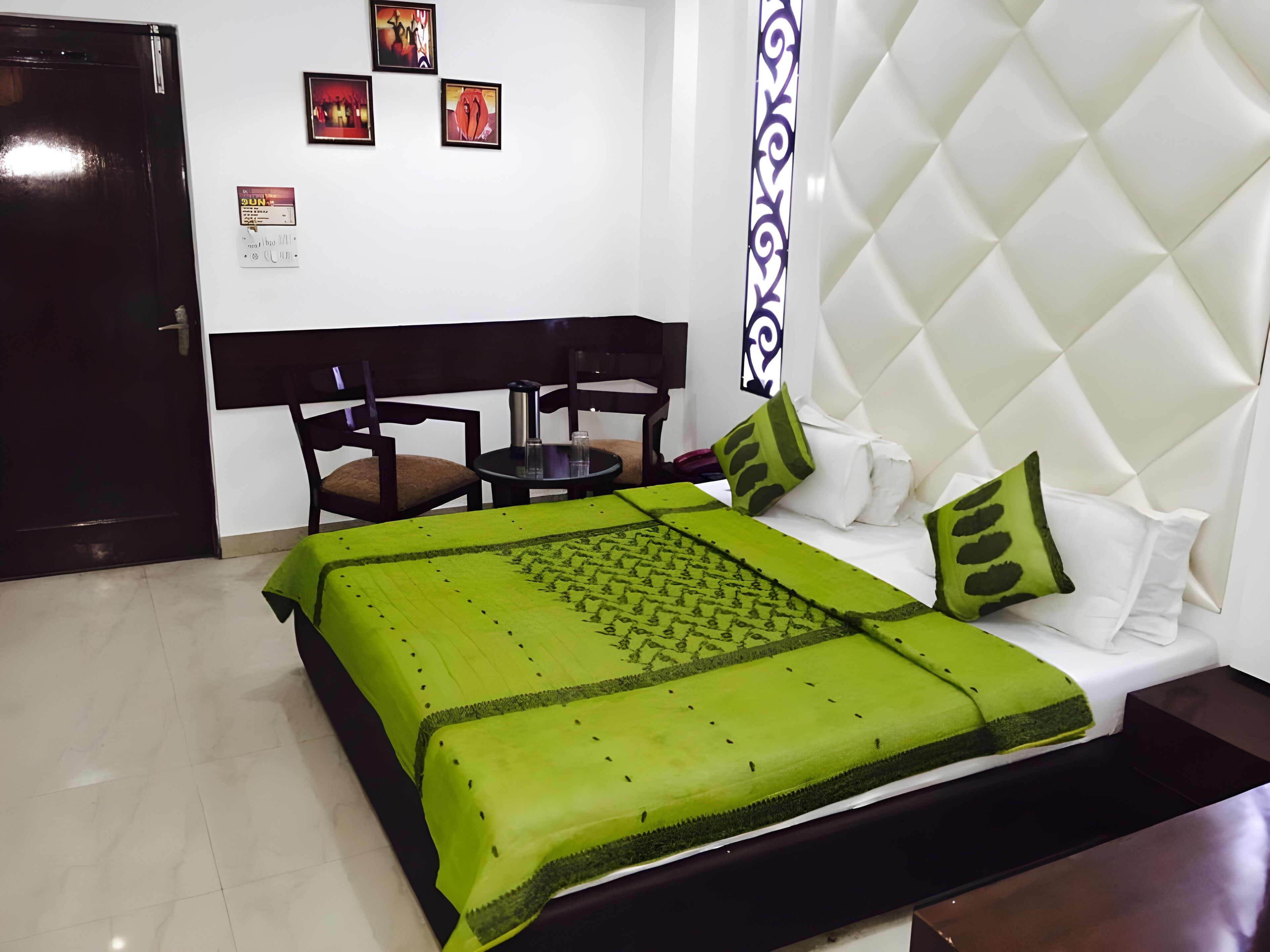 A hotel room with a bed, a small table, two chairs, a white headboard, and a door.